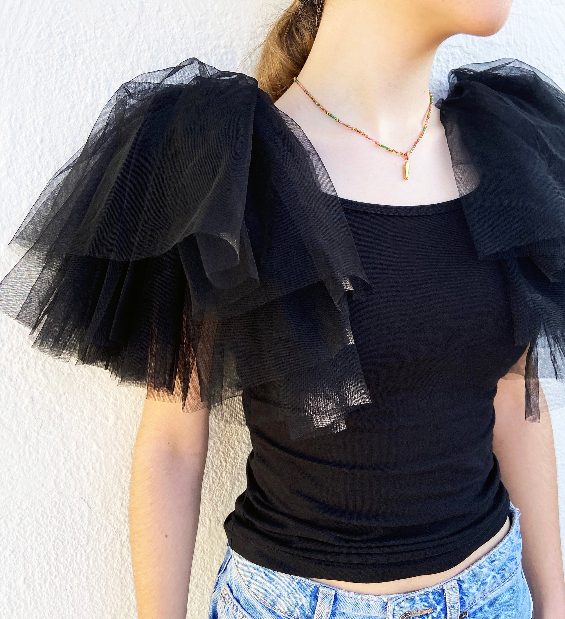 Black top with sheer tulle sleeves worn by a person against a white wall.
