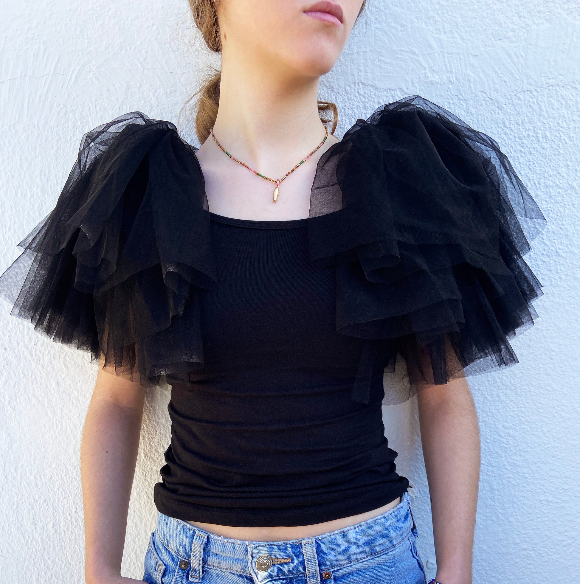 Black ruffled top with sheer sleeves worn by a person against a white wall.
