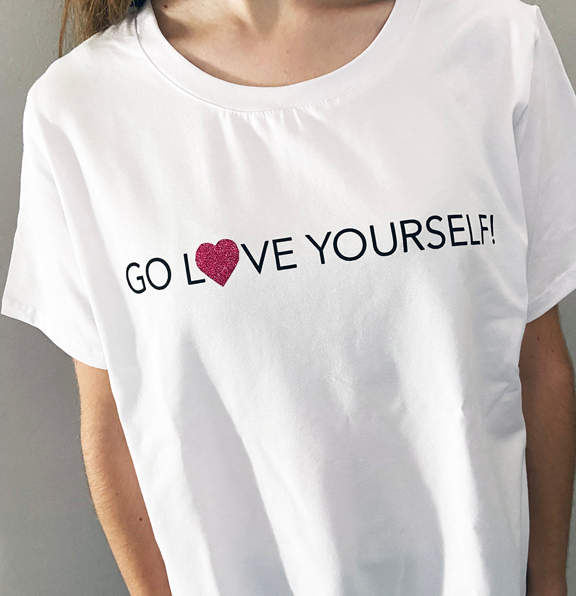 A close up of a person wearing a white t-shirt with the text 'GO LOVE YOURSELF!' and a glitter vinyl heart graphic on the front.