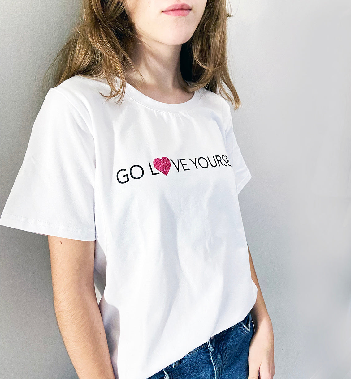 A person wearing a white t-shirt with the text 'GO LOVE YOURSELF!' and a glitter vinyl heart graphic on the front.