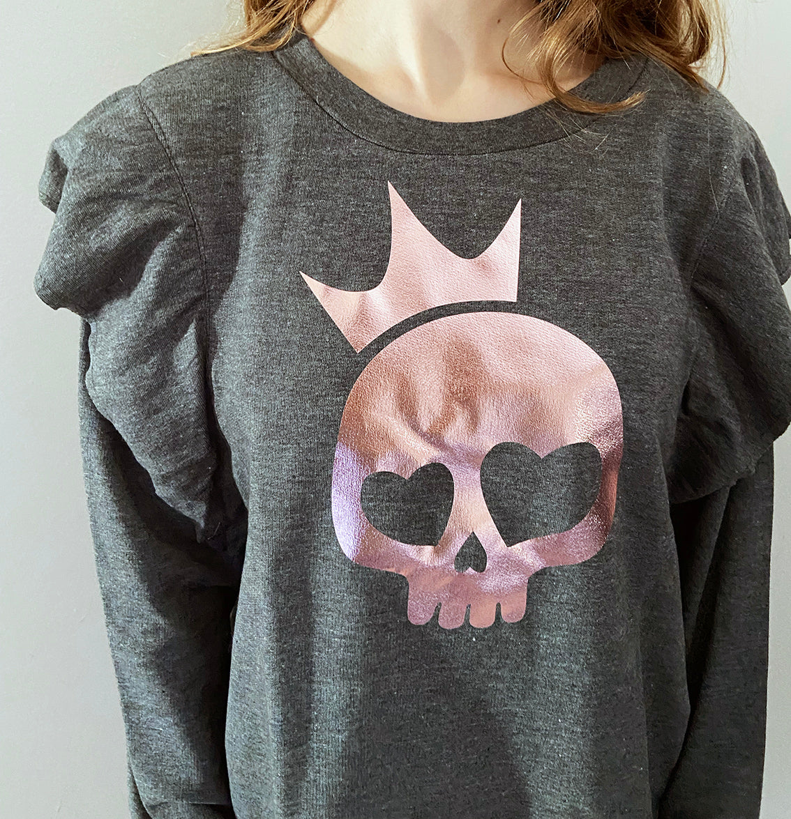 Close up of a person wearing a charcoal grey sweatshirt with a rose gold foil print of a skull with a crown design on the front.
