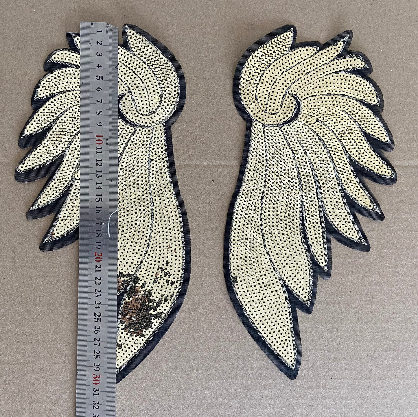 Two decorative wing-shaped patches with sequins on a brown background, accompanied by a ruler for scale.