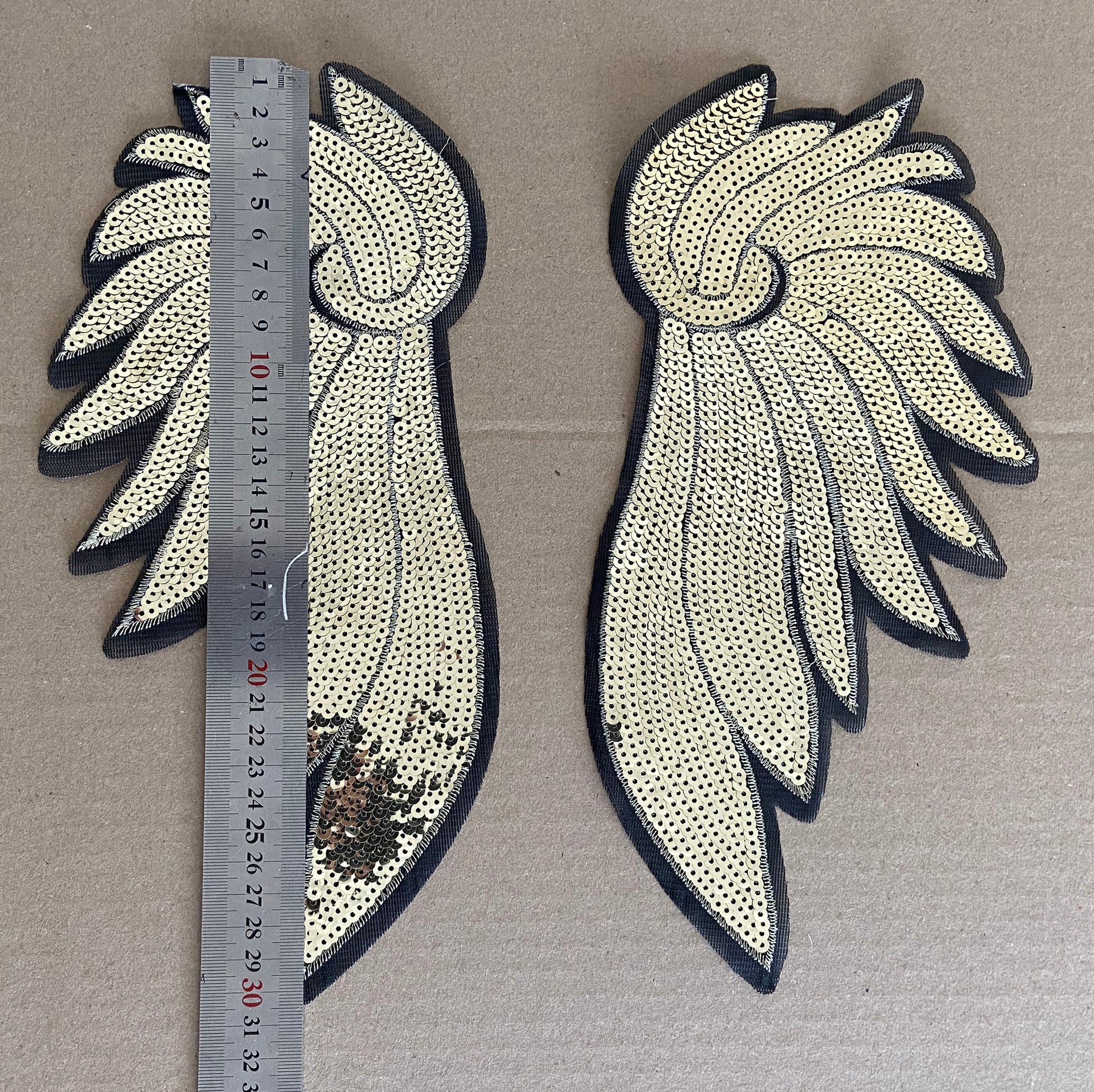 Two decorative wing-shaped patches with sequins on a brown background, accompanied by a ruler for scale.