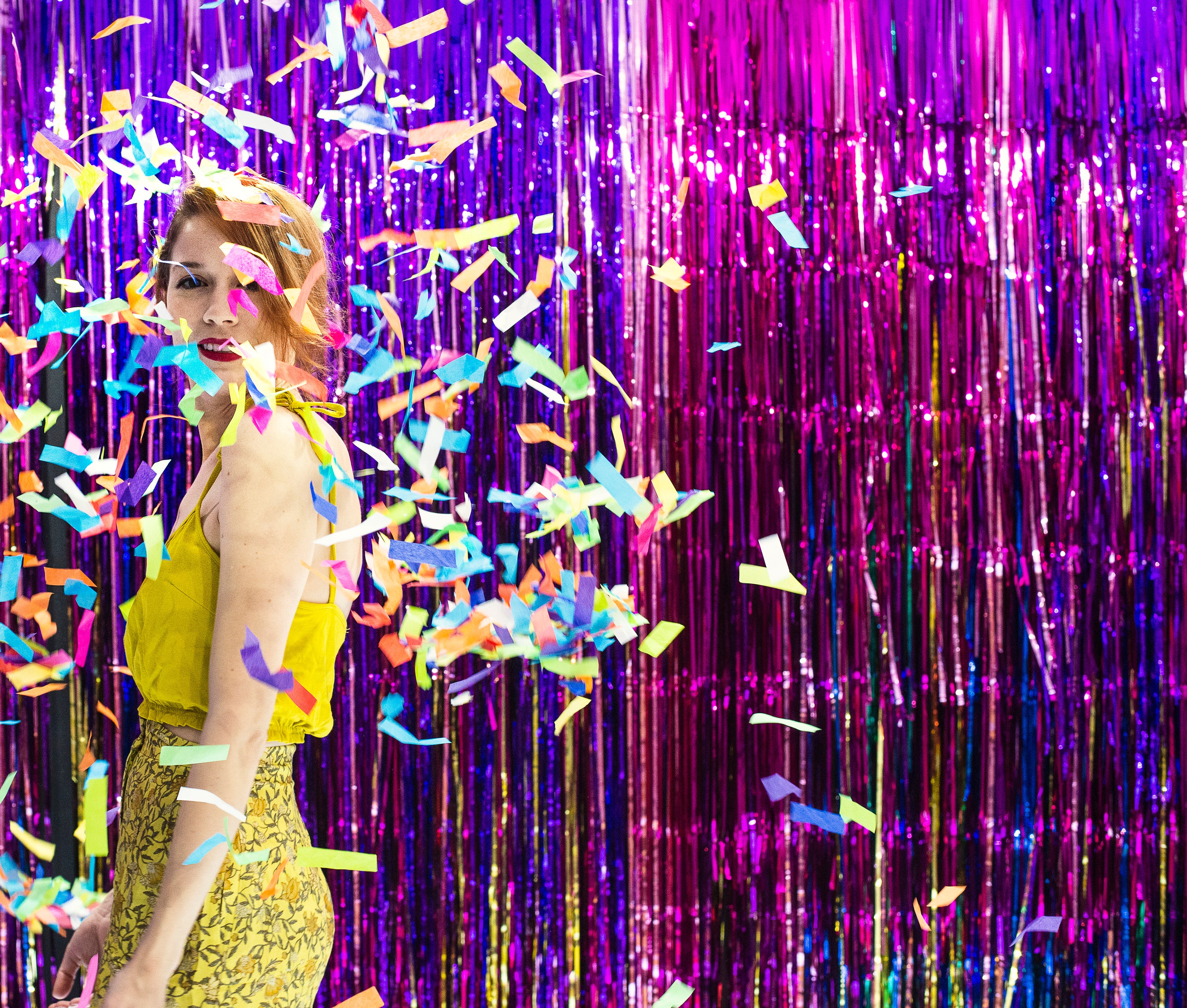 Woman in a yellow dress standing in front of a purple tinsel curtain with colorful confetti falling.