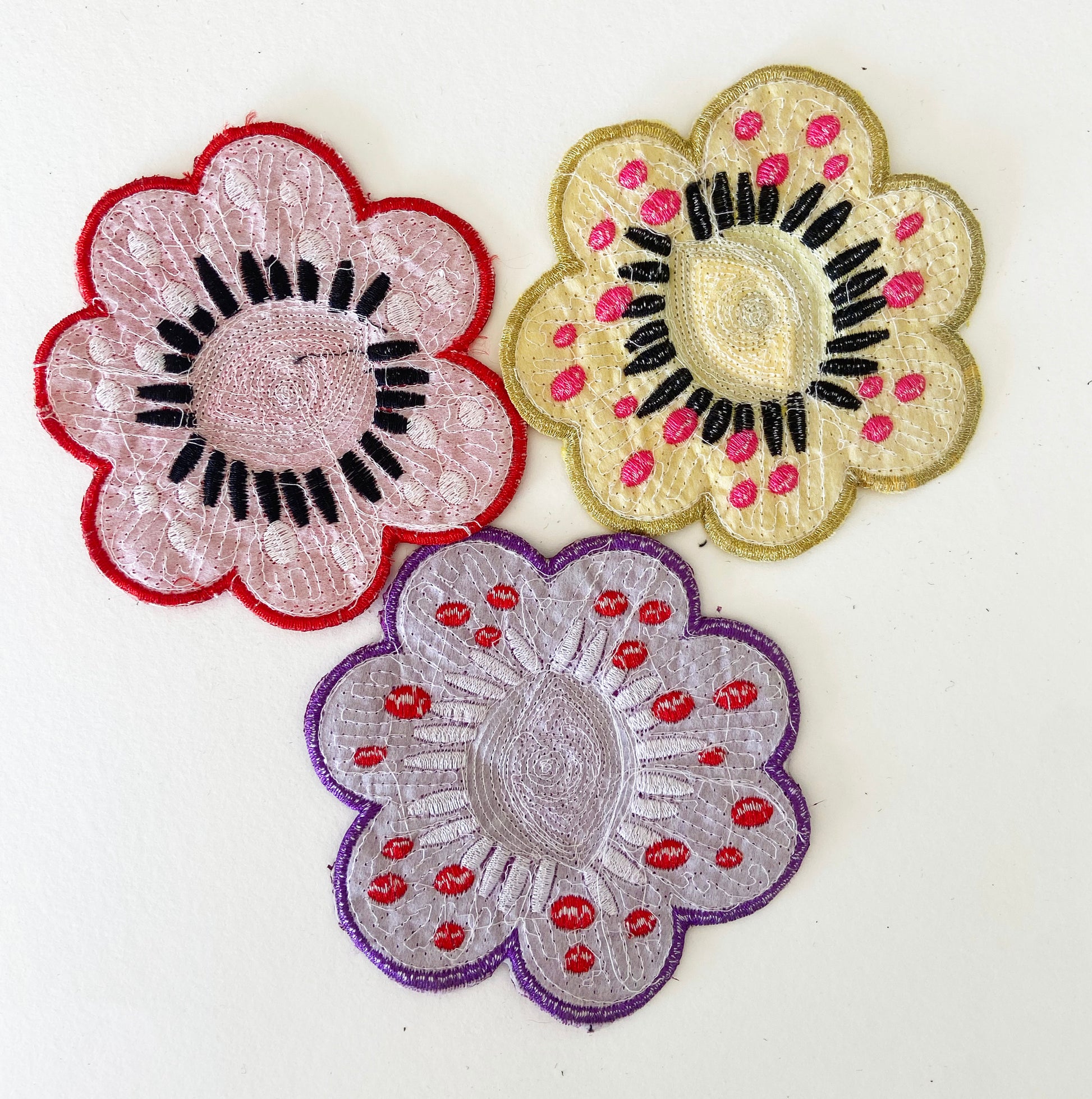 The backs of three embroidered flower patches in pink, gold, and purple on a white background