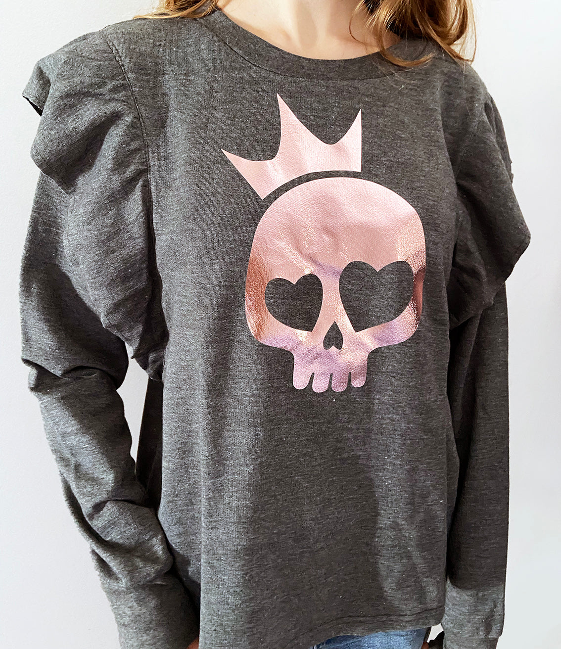 A person wearing a charcoal grey sweatshirt with a rose gold foil print of a skull with a crown design on the front.