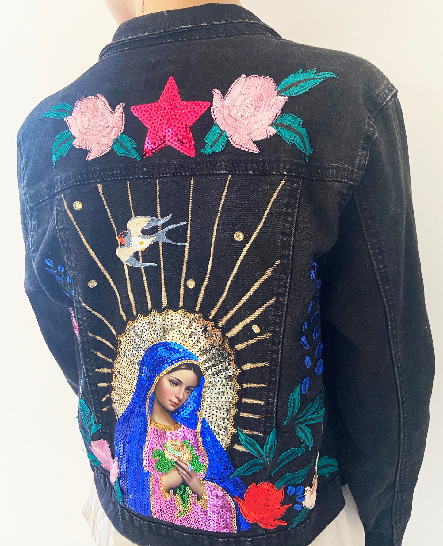 Black denim jacket with embroidered design of a religious figure, flowers, and stars.