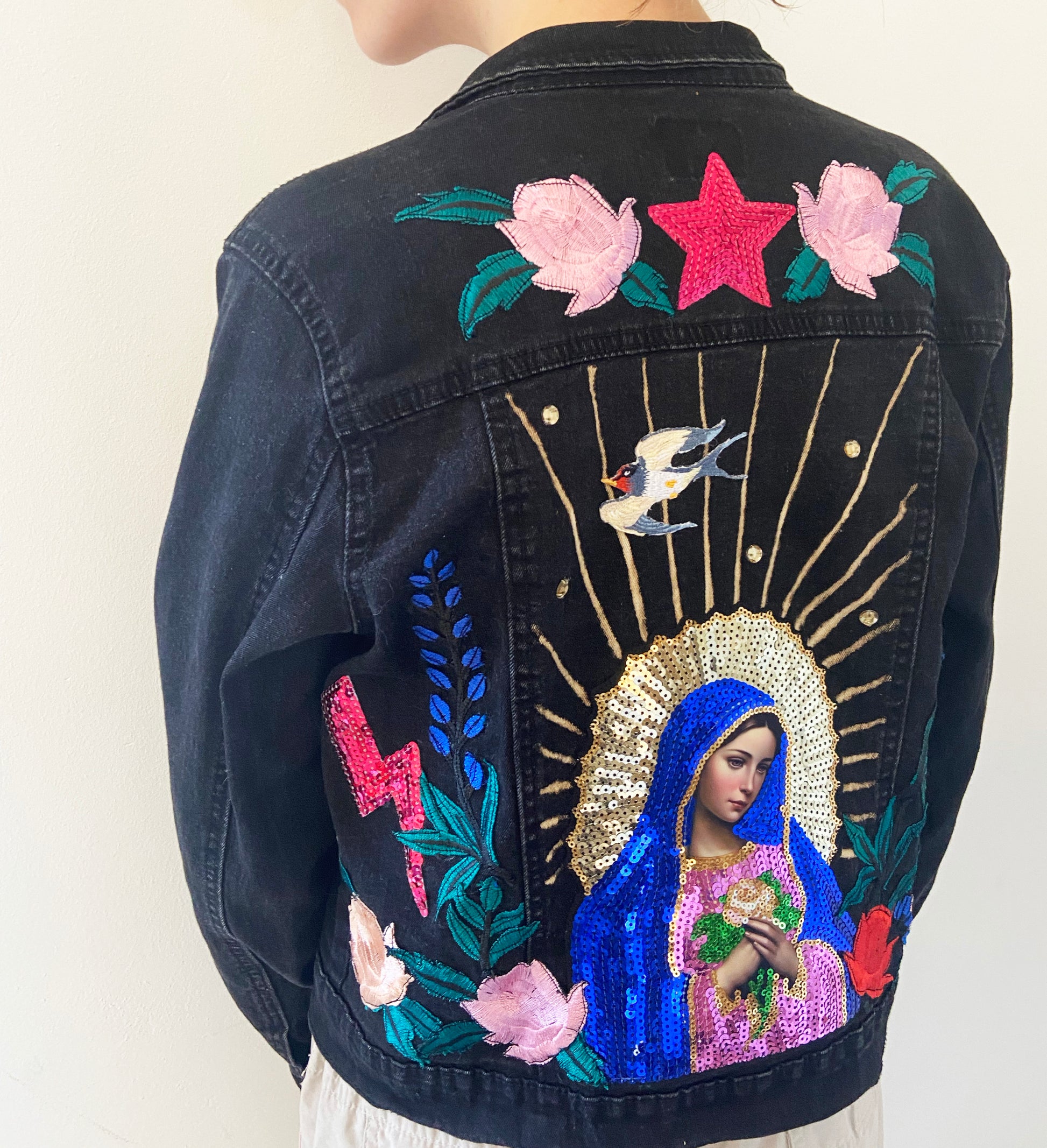 Black denim jacket with colorful embroidery of a religious figure, flowers, and a star.