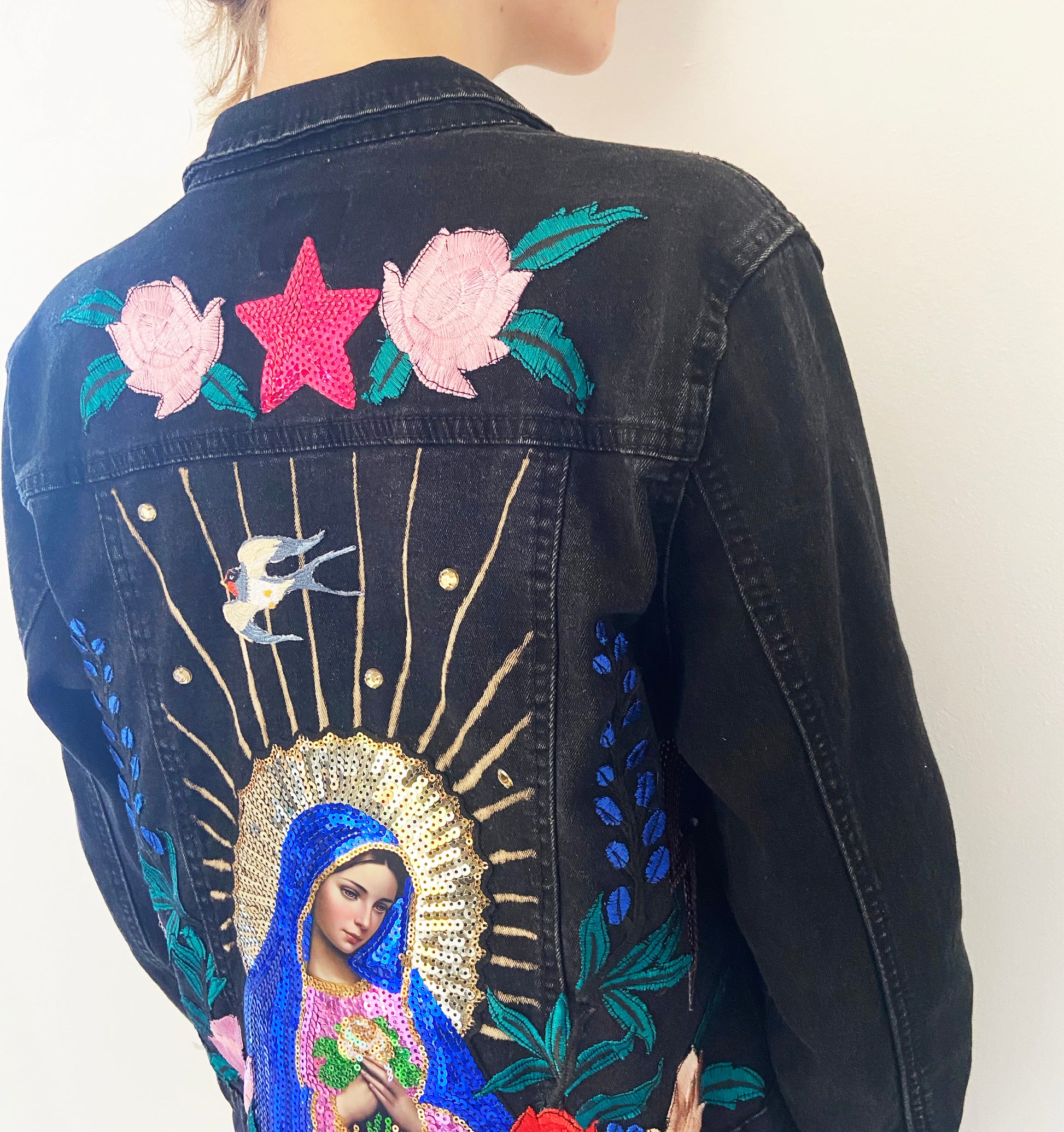 Black denim jacket with colorful embroidery of a religious figure, flowers, and stars.