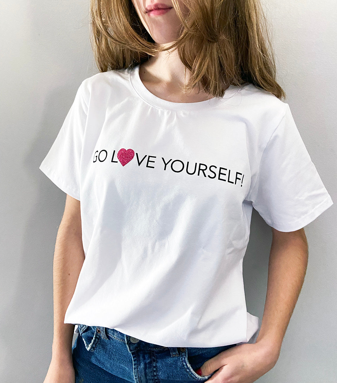 A close up of a person wearing a white t-shirt with the text 'GO LOVE YOURSELF!' and a glitter vinyl heart graphic on the front.