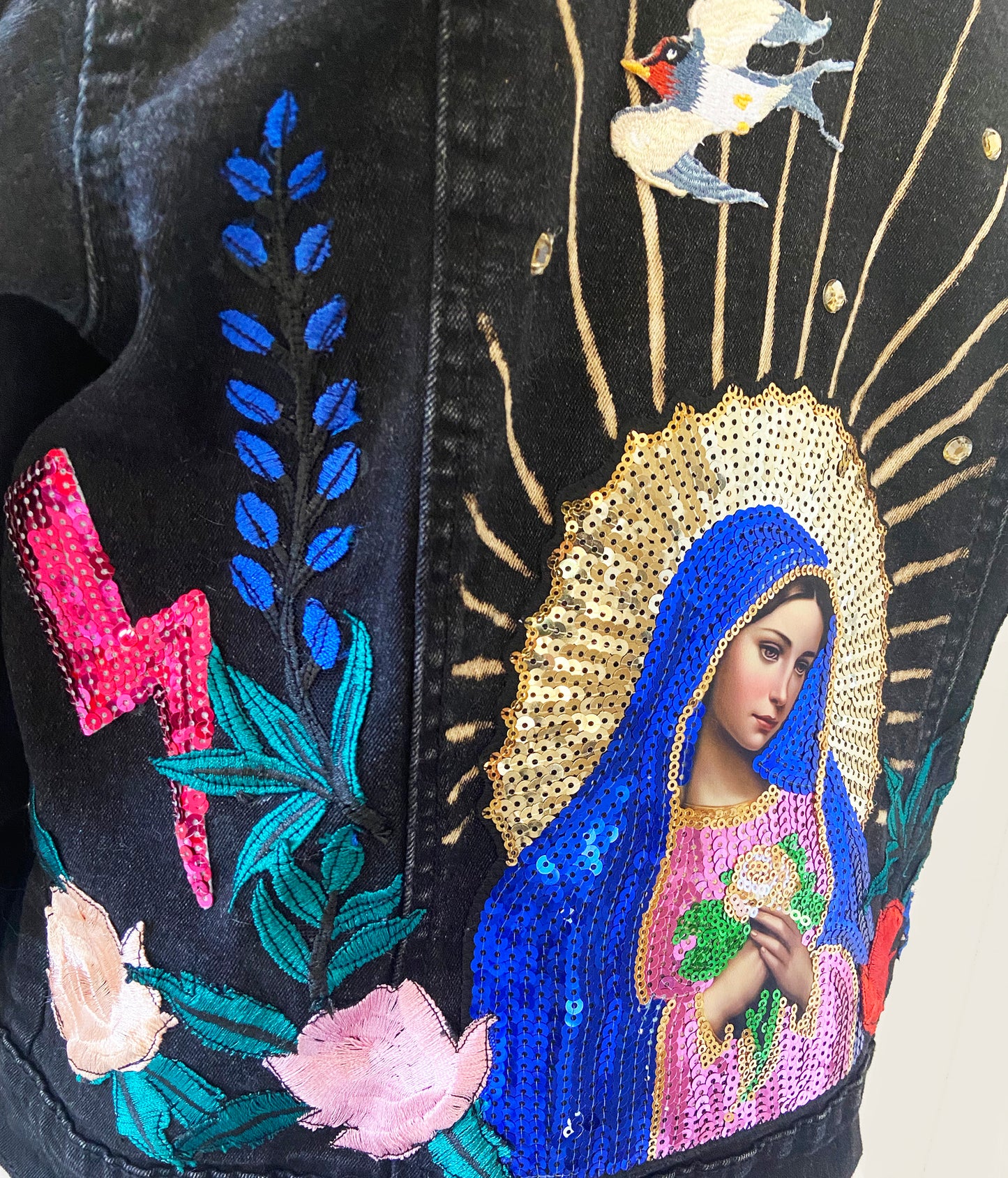 Embroidered design of a religious figure with flowers and a lightning bolt on a black background.