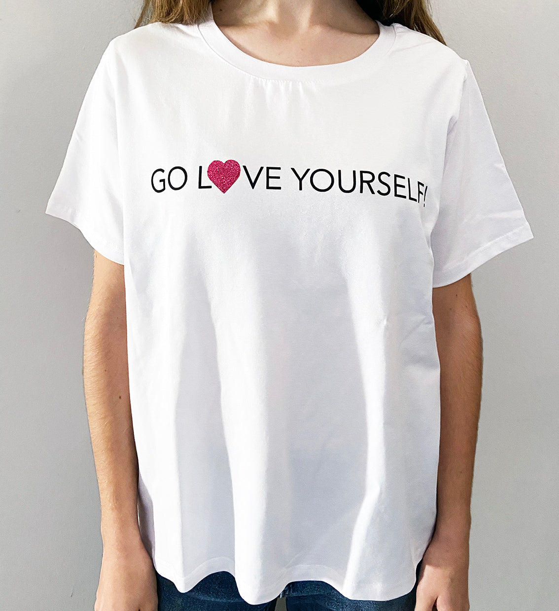 A person wearing a white t-shirt with the text 'GO LOVE YOURSELF!' and a glitter vinyl heart graphic on the front.