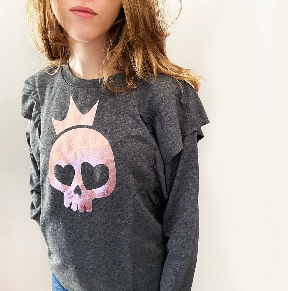 A person wearing a charcoal gray sweatshirt with a rose gold foil print of a skull with a crown design on the front.