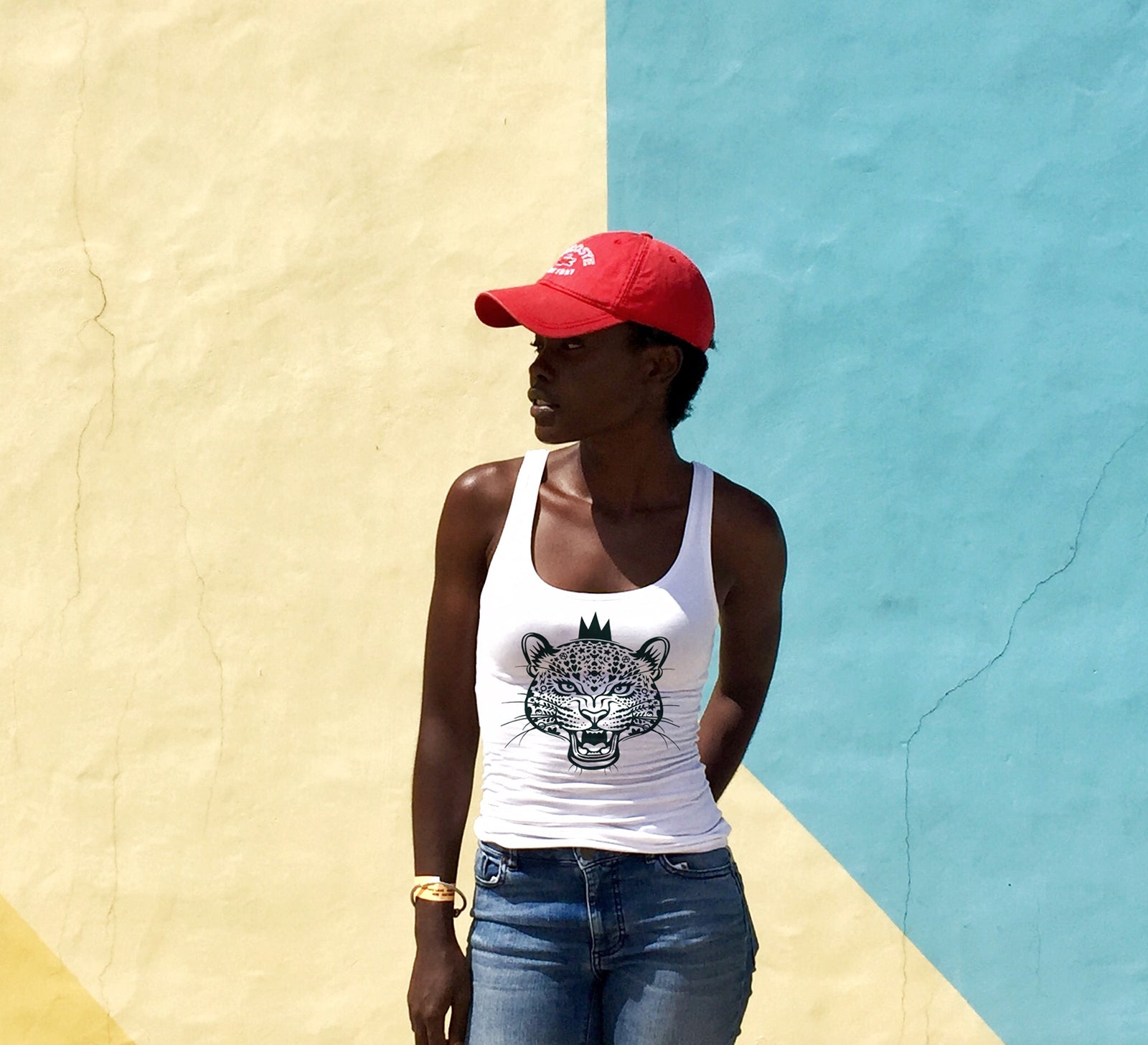 beautiful black woman wearing a red cap and white vest with green leopard queen print, standing in from of a brightly painted wall